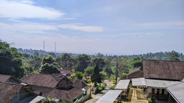 A rural landscape featuring a cluster of houses with tiled roofs surrounded by lush green trees. In the background, there are rolling hills and two visible communication towers. A narrow dirt road winds through the community, indicating a peaceful village setting under a partly cloudy sky.