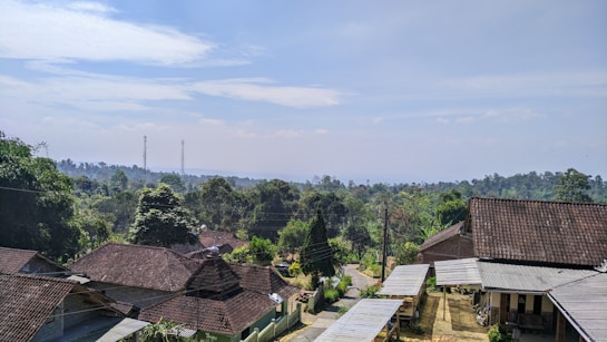 A rural landscape featuring a cluster of houses with tiled roofs surrounded by lush green trees. In the background, there are rolling hills and two visible communication towers. A narrow dirt road winds through the community, indicating a peaceful village setting under a partly cloudy sky.