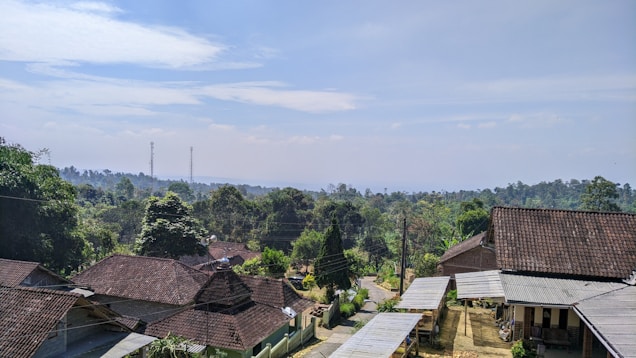 A rural landscape featuring a cluster of houses with tiled roofs surrounded by lush green trees. In the background, there are rolling hills and two visible communication towers. A narrow dirt road winds through the community, indicating a peaceful village setting under a partly cloudy sky.