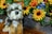 A plush toy dog with a fluffy, realistic appearance is sitting next to a wooden planter filled with vibrant artificial flowers, including sunflowers and various colorful blooms. The combination of textures from the soft fur and the vivid, lifelike flowers creates a cheerful and cozy scene.