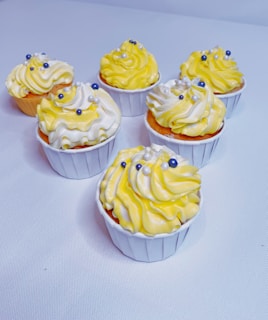Six cupcakes are arranged on a light surface, each topped with a swirl of yellow and white frosting. Blue and white candy pearls are delicately placed on top of the frosting as decoration.