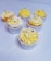 Six cupcakes are arranged on a light surface, each topped with a swirl of yellow and white frosting. Blue and white candy pearls are delicately placed on top of the frosting as decoration.