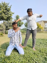 Two young men are outdoors in a park-like setting with trees and greenery. One man is kneeling on a patch of green plants wearing a striped shirt and blue jeans, while the other man, wearing a light-colored button-up shirt and dark pants, stands next to him with his hand posed over the kneeling man's head in a jovial manner. There is a building structure visible in the background.