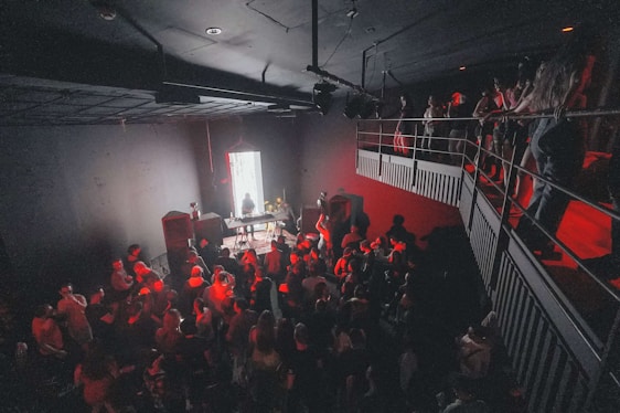 A dimly lit indoor space with a large crowd of people gathered around a DJ booth. The scene is illuminated with red lighting, and the room has an industrial feel with metal railings above. The DJ is positioned in front of a bright vertical light, creating a focal point in the image.