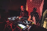 A DJ is performing with a mixing console, surrounded by technical equipment and glowing lights. The background features a large digital display with red patterns and a silhouetted shadow of the DJ.