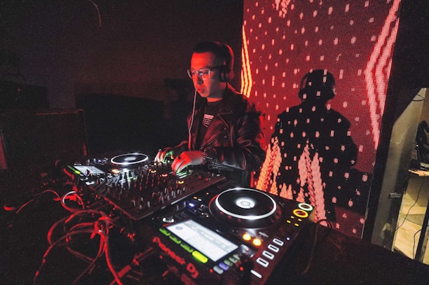 A DJ is performing with a mixing console, surrounded by technical equipment and glowing lights. The background features a large digital display with red patterns and a silhouetted shadow of the DJ.