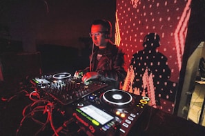 A DJ is performing with a mixing console, surrounded by technical equipment and glowing lights. The background features a large digital display with red patterns and a silhouetted shadow of the DJ.