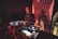 A DJ is performing with a mixing console, surrounded by technical equipment and glowing lights. The background features a large digital display with red patterns and a silhouetted shadow of the DJ.