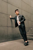 A model wearing a sleek black leather jacket paired with beige tapered pants, standing against a graffiti wall.