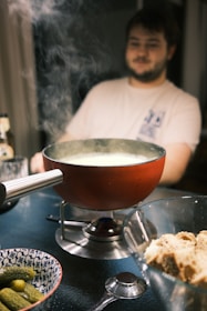 A person seated at a table with a fondue pot emitting steam, indicating it is hot and ready. A bowl with pickles and a glass dish containing chunks of bread are present on the table, suggesting a meal setting. The person appears relaxed and is watching the fondue pot.