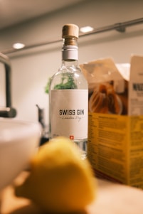 A bottle of Swiss gin labeled 'London Dry' is positioned on a kitchen counter. Nearby, there is a cardboard box with nutritional information, and a blurred lemon is in the foreground. The environment appears warmly lit, suggesting a cozy kitchen setting.