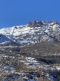 Majestic snow-capped mountains rise beneath a clear blue sky. Rugged peaks are adorned with snow, leading down to hilly terrain with sparse vegetation and patches of snow. The natural landscape is vast and untouched, providing a sense of peace and wilderness.