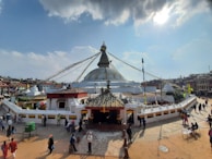 A large white stupa with a golden spire and eyes painted on it is surrounded by people walking and interacting in a square. Colorful prayer flags drape from the top of the stupa, and traditional buildings surround the area.