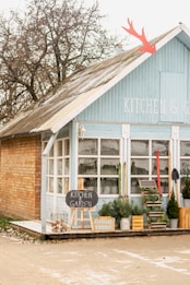 A quaint, blue painted building with white trim and a rustic roof. The signage reads 'KITCHEN & GARDEN' and there are wooden crates, potted plants, and a chalkboard sign at the entrance. The scene is complemented by a few red and green skis standing against the wall, a shovel, and bare-branched trees in the background, suggesting a winter setting.