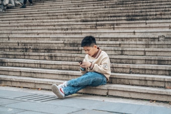 A child is sitting on a large set of concrete steps, absorbed in using a smartphone. The atmosphere is relaxed and casual, with the child wearing a light-colored sweater with design details and blue jeans. The setting is urban, with broad, evenly spaced steps visible in the background.