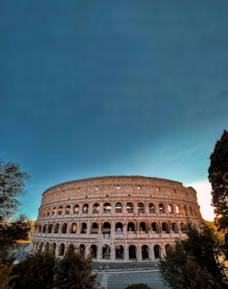 An impressive view of the Colosseum in Rome dominates the scene, with its ancient arches and stone structure illuminated by the warm light of the setting sun. Surrounding the historic amphitheater are scattered trees and a clear, expansive sky.