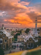 A stunning view of a cityscape at sunset, featuring unique, whimsical architecture characteristic of Gaudí's style in the foreground. The sky is ablaze with vibrant orange and yellow hues, creating a dramatic backdrop against the city buildings.