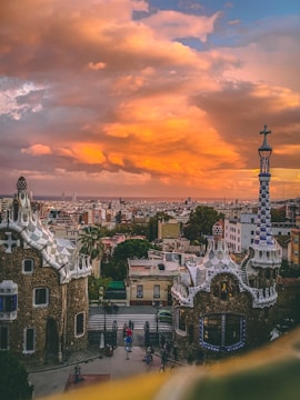 A stunning view of a cityscape at sunset, featuring unique, whimsical architecture characteristic of Gaudí's style in the foreground. The sky is ablaze with vibrant orange and yellow hues, creating a dramatic backdrop against the city buildings.