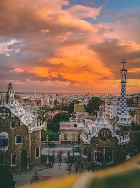 A stunning view of a cityscape at sunset, featuring unique, whimsical architecture characteristic of Gaud&iacute;'s style in the foreground. The sky is ablaze with vibrant orange and yellow hues, creating a dramatic backdrop against the city buildings.