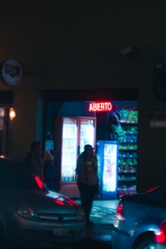 A nighttime street scene with a small store, illuminated by a red neon sign that reads 'ABIERT0.' The store features refrigerated displays and snack racks inside, with a few people standing near the entrance. Cars are parked on the street, reflecting the neon lights.