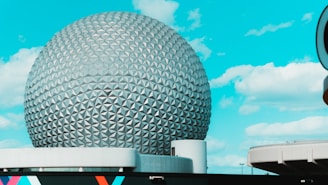 A large geodesic sphere structure with a pattern of triangular panels is set against a bright blue sky with scattered clouds. The base of the structure has modern architectural elements and a segment of a building is visible in the foreground.