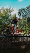 A theme park attraction resembling a mountain with a waterfall. Lush greenery surrounds the structure, while a wooden bridge with people walking across it is visible in the foreground.