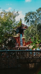A theme park attraction resembling a mountain with a waterfall. Lush greenery surrounds the structure, while a wooden bridge with people walking across it is visible in the foreground.
