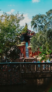 A theme park attraction resembling a mountain with a waterfall. Lush greenery surrounds the structure, while a wooden bridge with people walking across it is visible in the foreground.