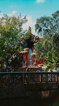 A theme park attraction resembling a mountain with a waterfall. Lush greenery surrounds the structure, while a wooden bridge with people walking across it is visible in the foreground.