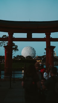 A torii gate with a spherical structure in the background, likely resembling Epcot's Spaceship Earth. The scene is set during dusk with a crowd of people gathered around, trees, and a body of water also visible.