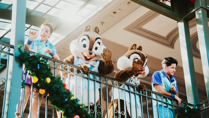 Two people in costumes resembling animated characters wave from an elevated platform alongside two adults dressed in blue uniforms. A decorative garland with colorful ornaments adorns the railing.
