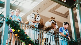 Two people in costumes resembling animated characters wave from an elevated platform alongside two adults dressed in blue uniforms. A decorative garland with colorful ornaments adorns the railing.