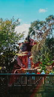 A theme park attraction with a waterfall cascading down a rocky structure surrounded by lush trees. People can be seen gathered on a wooden bridge in the foreground, looking towards the ride.