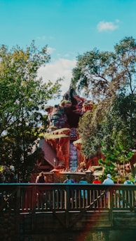 A theme park attraction with a waterfall cascading down a rocky structure surrounded by lush trees. People can be seen gathered on a wooden bridge in the foreground, looking towards the ride.