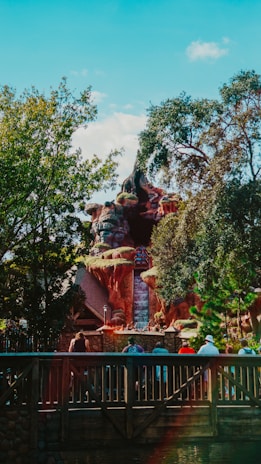 A theme park attraction with a waterfall cascading down a rocky structure surrounded by lush trees. People can be seen gathered on a wooden bridge in the foreground, looking towards the ride.