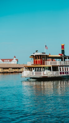 A riverboat cruises on a wide expanse of calm water, featuring an American flag and a nameplate reading 'Richard F. Irvine'. The sky is clear and blue, and in the background, there is a building with a red roof.