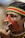 A person with traditional face paint and tattoos is seen holding a wooden object to their nose. They are wearing a colorful headband with red, yellow, and green stripes and their attire includes intricate beads. Their ear is adorned with a large earring.