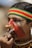 A person with traditional face paint and tattoos is seen holding a wooden object to their nose. They are wearing a colorful headband with red, yellow, and green stripes and their attire includes intricate beads. Their ear is adorned with a large earring.