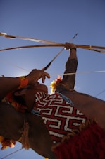 A person dressed in traditional attire is aiming a bow and arrow towards the sky. The image is captured from a low angle, highlighting the arm muscles and the intricate beadwork on the clothing. The bright blue sky forms the backdrop, creating a dramatic composition that focuses on the action and the cultural elements.