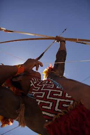 A person dressed in traditional attire is aiming a bow and arrow towards the sky. The image is captured from a low angle, highlighting the arm muscles and the intricate beadwork on the clothing. The bright blue sky forms the backdrop, creating a dramatic composition that focuses on the action and the cultural elements.