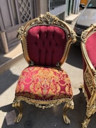 An ornate chair featuring a rich burgundy and gold floral upholstery with gold-painted wooden frame and intricate carvings. It is positioned outdoors on a street, next to another matching chair.