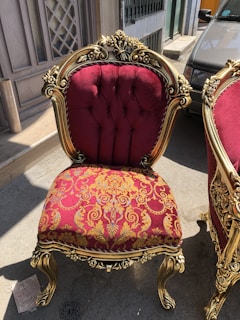 An ornate chair featuring a rich burgundy and gold floral upholstery with gold-painted wooden frame and intricate carvings. It is positioned outdoors on a street, next to another matching chair.