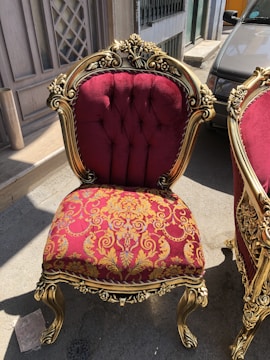 An ornate chair featuring a rich burgundy and gold floral upholstery with gold-painted wooden frame and intricate carvings. It is positioned outdoors on a street, next to another matching chair.