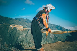 A person wearing traditional attire with a colorful headscarf and woven garment stands on a mountainous landscape. They are holding a bow-like object and appear focused on their task. The background features lush rolling hills under a clear blue sky.