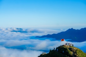 A group of people stand on a mountain peak with a Vietnamese flag against a backdrop of rolling clouds. The scene is set against expansive blue sky and distant mountains shrouded in mist, creating a sense of vastness and elevation.