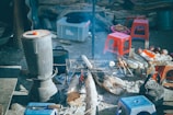 Various cooking utensils and ingredients are arranged around an outdoor campfire. There is a grill with skewered foods cooking over the fire, a large pot on a stand, and several small red and blue plastic stools. A plastic storage box and some wooden logs can be seen in the background.