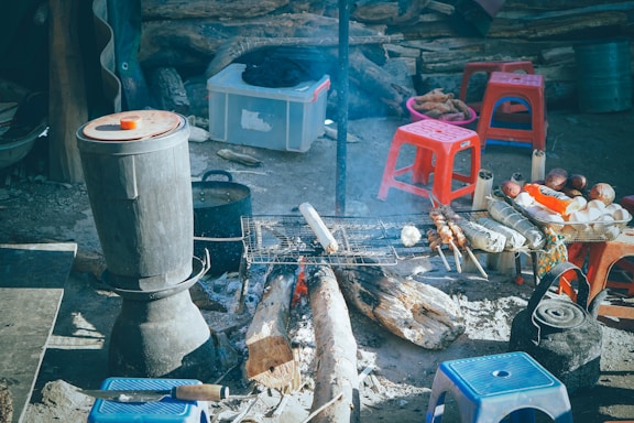 Various cooking utensils and ingredients are arranged around an outdoor campfire. There is a grill with skewered foods cooking over the fire, a large pot on a stand, and several small red and blue plastic stools. A plastic storage box and some wooden logs can be seen in the background.
