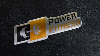 A metal plaque with the text 'POWER FITNESS' is set against a textured black background. The design includes a stylized dumbbell icon adjacent to the text, with a modern and sleek appearance.