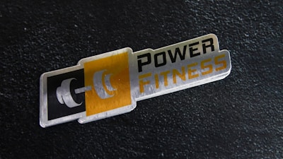 A metal plaque with the text 'POWER FITNESS' is set against a textured black background. The design includes a stylized dumbbell icon adjacent to the text, with a modern and sleek appearance.