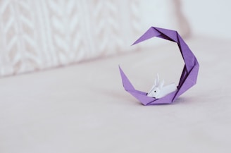 An origami sculpture featuring a small white rabbit resting within a crescent-shaped purple structure. The background is an off-white, textured surface that complements the delicate and intricate design of the origami.